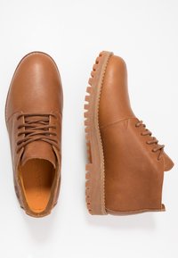 Botas de cuero marrón con una textura suave, puntera redonda y suela resistente con un diseño de tacos distintivo. Cordones y costuras visibles en los laterales.