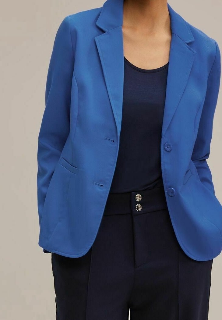 Blazer bleu en tissu lisse, avec des revers à encoches, deux boutons et des poches latérales, porté avec un haut sombre et un pantalon taille haute.