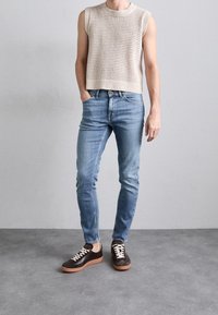 Beige stickad ärmlös topp, blå slim-fit jeans och mörkbruna skor med vita snörningar och gummisålar. Slät textur och avslappnad design.