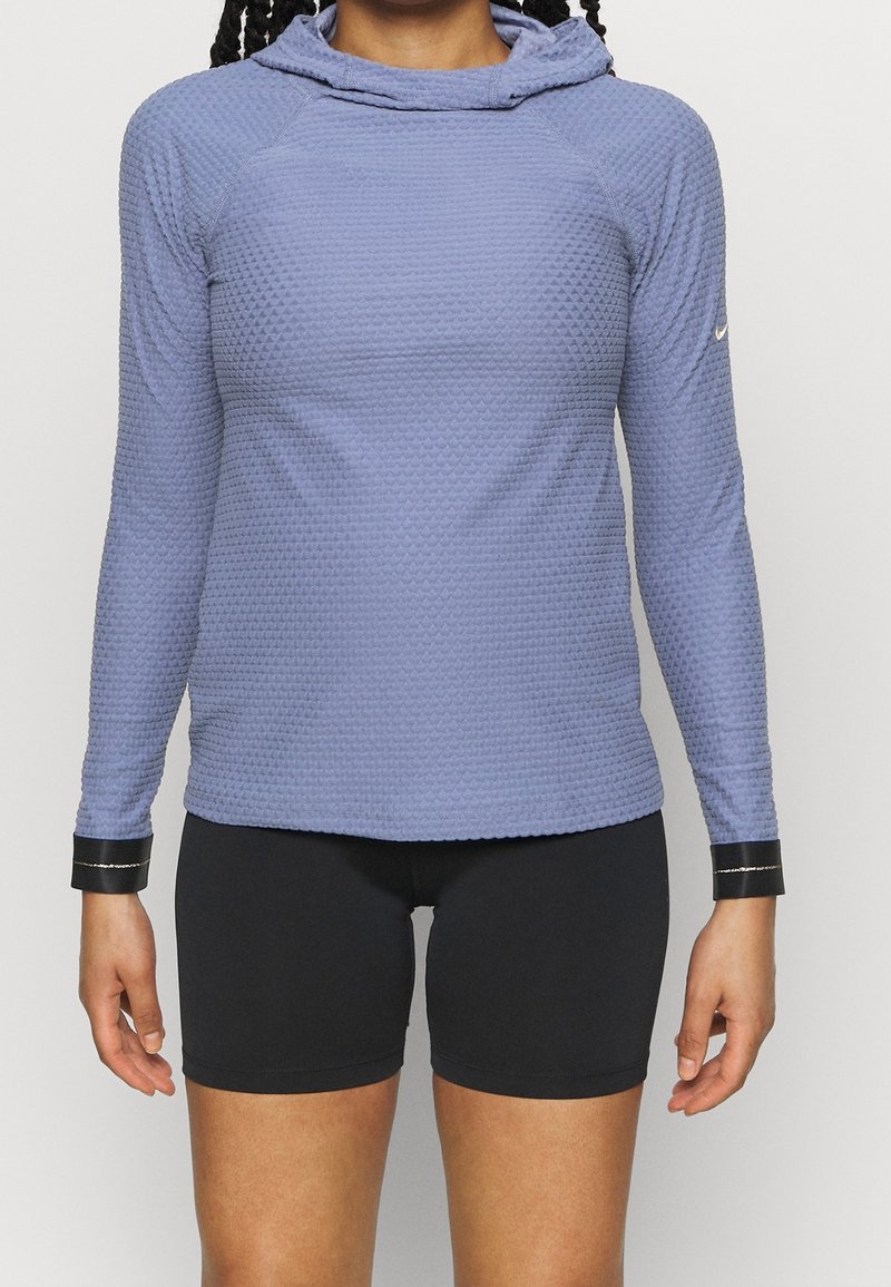 Femme portant un haut à capuche texturé bleu à manches longues et un short noir ajusté, debout, les bras détendus le long du corps.