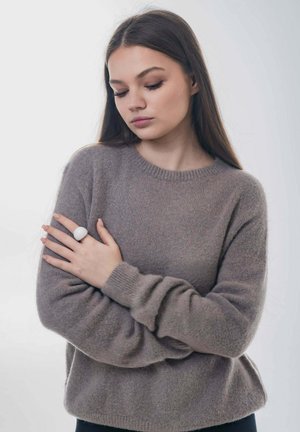 Młoda kobieta o długich brązowych włosach, ubrana w szary dzianinowy sweter, z założonymi skrzyżowanymi ramionami, patrzy w dół z zamkniętymi oczami w miękkim oświetleniu.