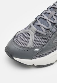 Grå sneakers med övre av mesh, vadderad tunga och en texturerad gummisula. Har detaljer i mörkare grått och ljusgrå accenter.