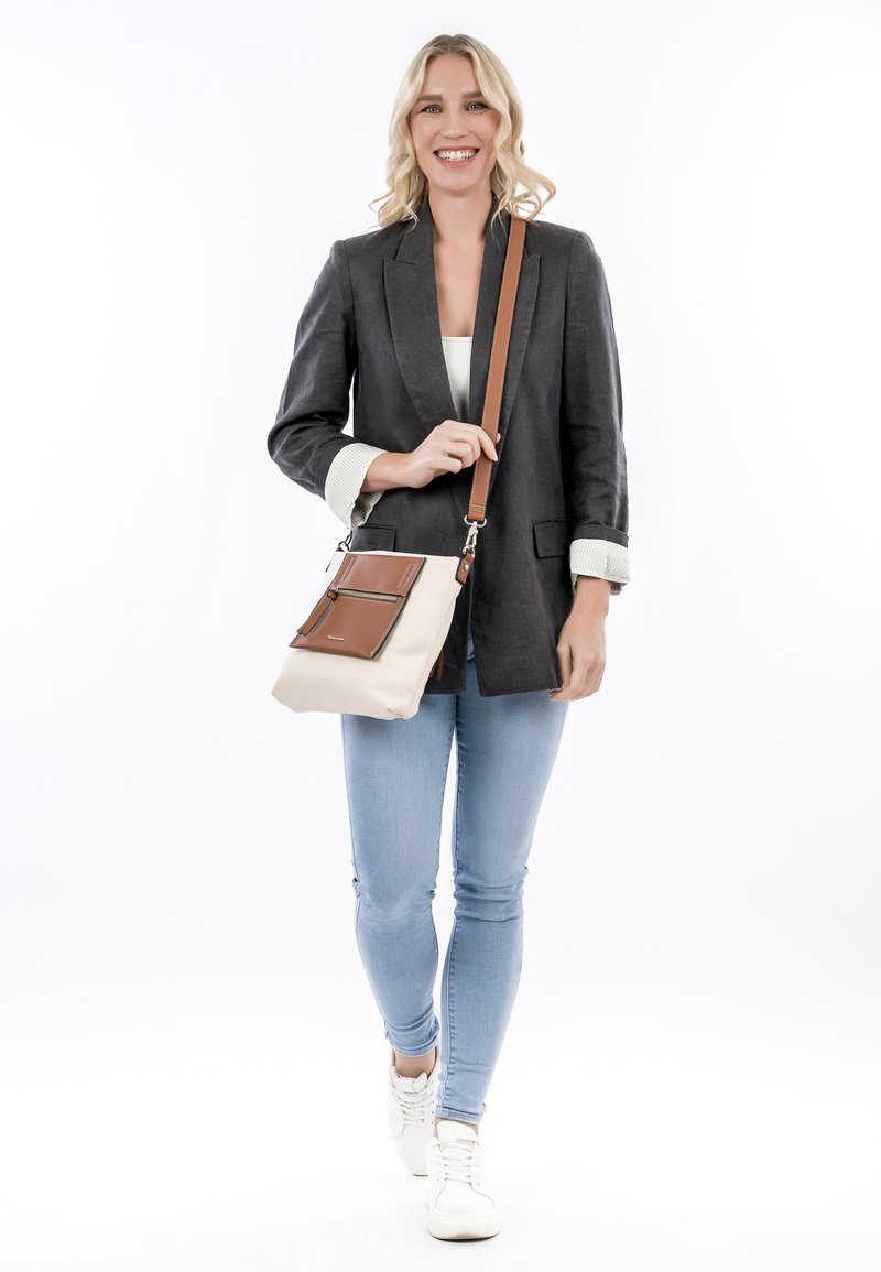 Hnědo-krémová crossbody taška s hladkým povrchem, dvěma předními kapsami a nastavitelným popruhem. Modelka má na sobě šedou blazer a džíny.