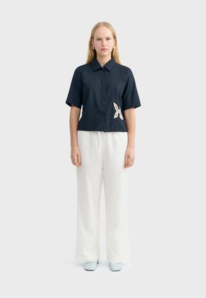 Blondínka stojaca, oblečená v čiernej krátkorevej košeli na gombíky s bielou záplatou v tvare hviezdice a bielych nohaviciach so širokými nohavicami.