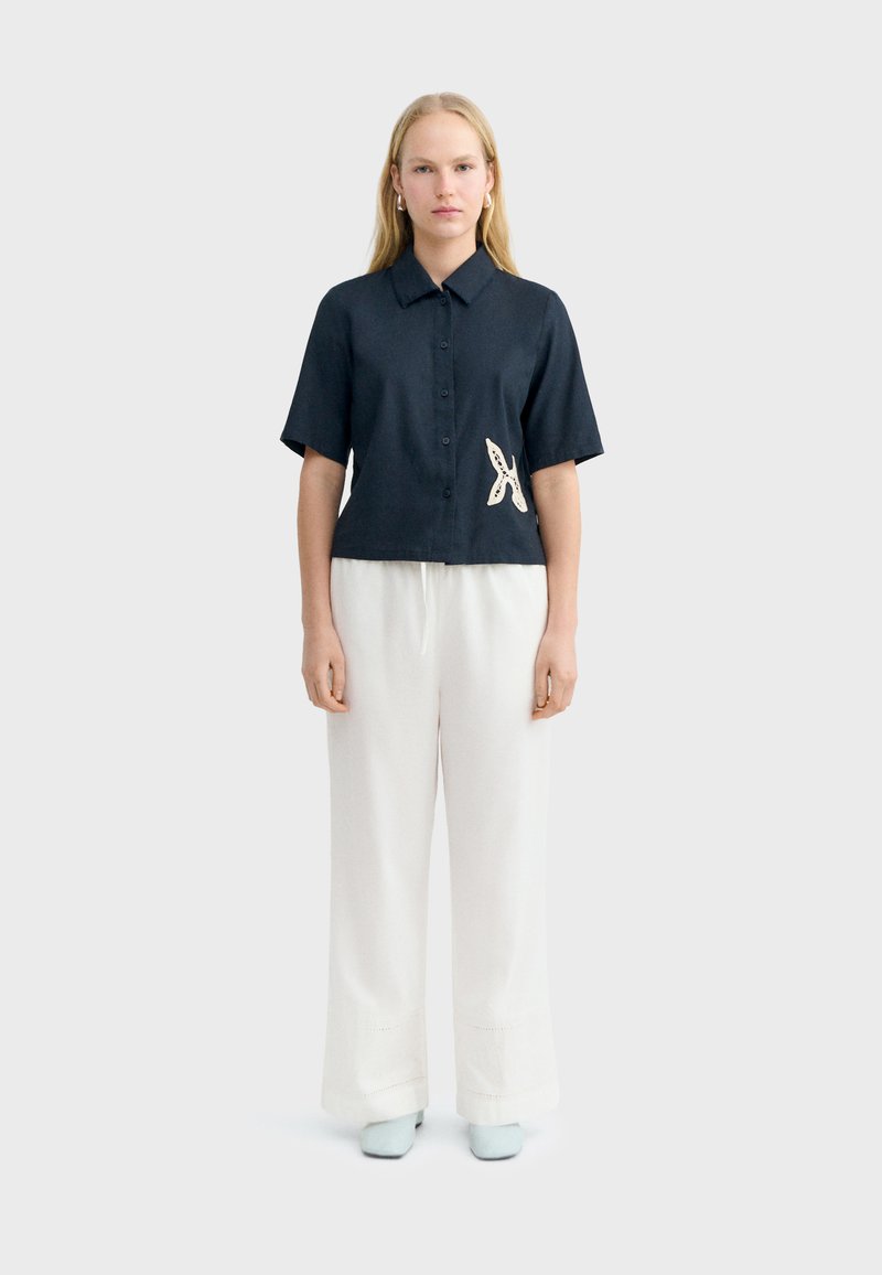 Femme blonde debout, portant une chemise noire à manches courtes avec un patch d'étoile de mer blanche et un pantalon blanc à jambes larges.