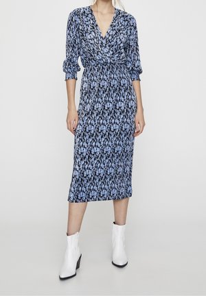 Vrouw die een blauwe en zwarte midi-jurk met bloemenprint en driekwartmouwen draagt, samen met witte enkellaarzen, staand tegen een effen lichte achtergrond.