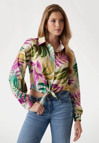 Overhemd met knopen, versierd met levendige groene en paarse bladeren op een lichtgele achtergrond; geknoopt aan de voorkant; lange mouwen; gecombineerd met blauwe jeans.