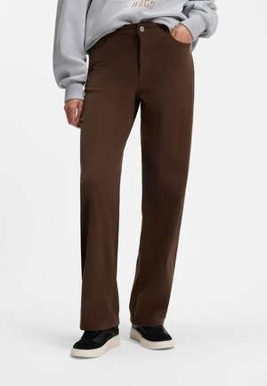Person, die braune gerade geschnittene Hosen, schwarz-weiße Sneakers und ein graues Sweatshirt mit Text trägt, steht mit einer Hand in der Tasche.