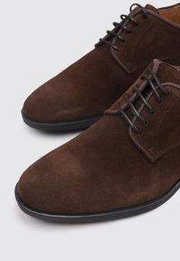 Zapatos de vestir de ante marrón con una textura suave, punta redonda y diseño de cordones. Cuentan con finas suelas de goma negra y sutiles acentos de costura.