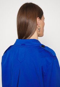 Femme aux cheveux bruns et raides, portant une veste bleue avec "BOSS" brodé sur le col et des boucles d'oreilles argentées en forme d'anneau, tournée de dos à la caméra.