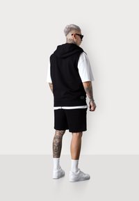 Schwarzer ärmelloser Hoodie über einem weißen T-Shirt, kombiniert mit schwarzen Shorts. Weiße Sneaker und sichtbare Tattoos am Bein. Einfaches Design.