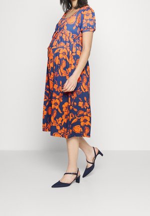 Umstandskleid mit marineblauem Hintergrund und großen orangefarbenen Blumenmustern, kurzen Ärmeln und einem ausgestellten Schnitt. Kombiniert mit marineblauen Pumps.