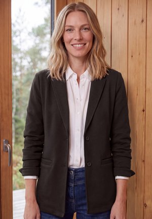 Mujer rubia sonriente que lleva una blazer negra, camisa blanca y vaqueros azules, de pie en el interior cerca de una pared de madera y una ventana con vegetación exterior visible.