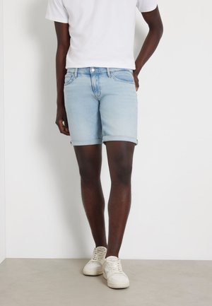 Shorts de mezclilla azul claro con dobladillos enrollados, que presentan un diseño clásico de cinco bolsillos y cierre de botón de metal, combinados con una camiseta blanca y zapatillas deportivas.
