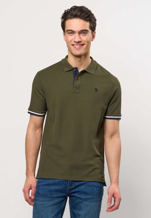 Noor mees naeratab, kannab oliivirohelist polosärki tumeda äärega varrukatel ja krae juures, siniste teksadega, seisab ühevärvilise tausta ees.