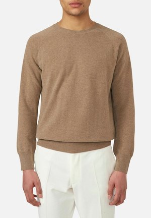Uomo che indossa un maglione girocollo marrone chiaro con maniche lunghe e pantaloni bianchi plissettati, in piedi davanti a uno sfondo semplice.