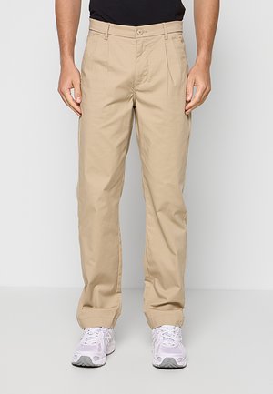 Mann trägt beige gerade geschnittene Hose mit Bundfalten, weiße Sneakers und schwarzes Hemd, steht vor hellem grauem Hintergrund.