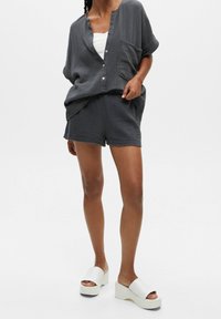 Chemise grise texturée avec une poche, assortie à un short. La tenue est complétée par des sandales compensées blanches.