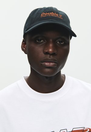 Młoda osoba o ciemnej karnacji, nosząca czarną czapkę z pomarańczowym napisem „Double V” oraz białą koszulkę z częściowo widoczną grafiką, o neutralnym wyrazie twarzy.