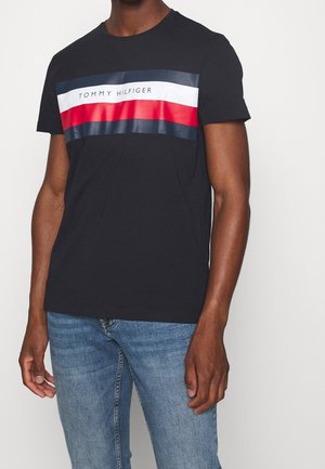 Homme portant un t-shirt noir Tommy Hilfiger avec des bandes horizontales rouges, blanches et bleues sur la poitrine et un jean bleu.