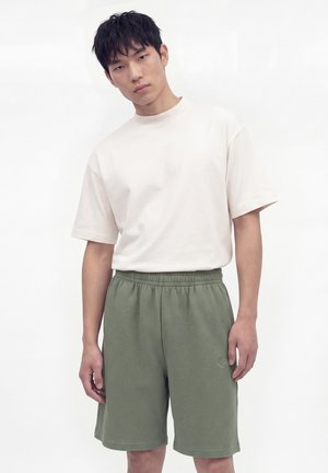 Jeune homme aux cheveux noirs et courts portant un T-shirt blanc ample et un short vert, debout devant un fond blanc uni, regardant vers la caméra.