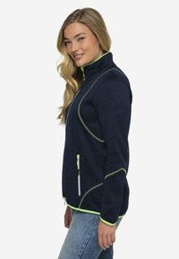 Giacca blu navy con zip e colletto alto, caratterizzata da dettagli verde lime, cuciture curve e tasche con zip. Tessuto testurizzato.
