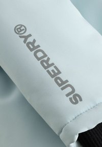 Manche en tissu bleu clair avec le logo "Superdry" gris imprimé en diagonale et un poignet élastique noir visible au bord inférieur.