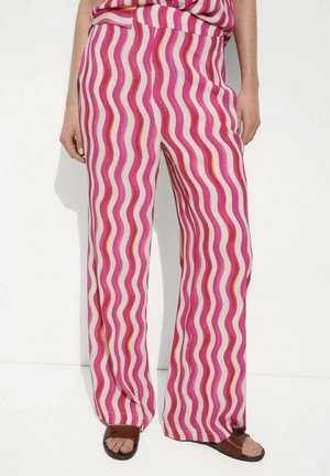 Mujer con pantalones de talle alto y pierna ancha con rayas onduladas verticales en rosa, blanco y naranja, combinados con sandalias marrones de punta abierta.