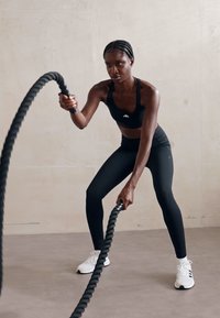Fekete sportos nő fekete crop topban és leggingsben vastag fekete küzdőköteleket használ. Fehér sportcipőt visel fekete részletekkel.