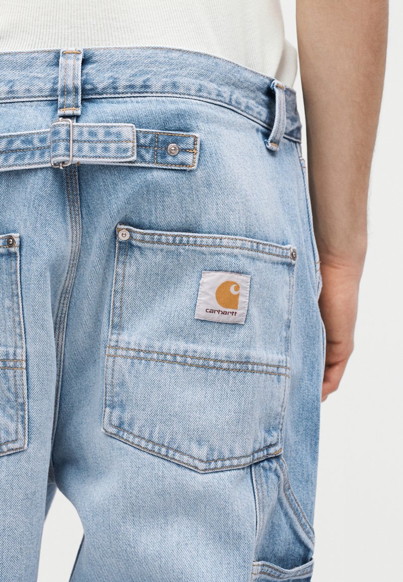 Jeans in denim azzurro chiaro con tasca posteriore che presenta una toppa con il logo Carhartt e cinturino regolabile in vita, indossati da una persona con maglietta bianca.