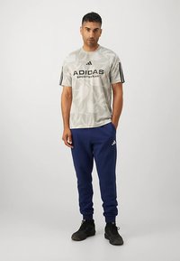 Pilkas Adidas sportinis marškinėlis su juodu logotipu ir šoninėmis juostelėmis, derinamas su mėlynomis sportinėmis kelnėmis su Adidas logotipu ant kojos.