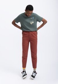Donker groene katoenen t-shirt met een grafisch ontwerp; roestkleurige tapered broek; zwarte canvas sneakers met witte zolen en sokken met gele accenten.