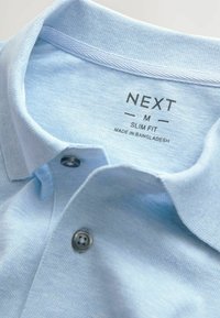 Ljusblå pikétröja med texturerad tyg, slim fit-design och tvåknappsslå. Etiketten säger "NEXT M SLIM FIT MADE IN BANGLADESH."