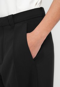 Pantalon noir à coupe droite avec poches latérales ; texture lisse et coupe ajustée ; la ceinture est équipée de passants pour ceinture.