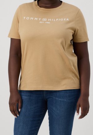 Kobieta w beżowej koszulce Tommy Hilfiger z logo i napisem "EST. 1985", w niebieskich dżinsach, stojąca na jednolitym tle.