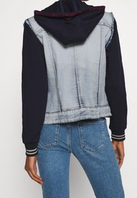 Veste bomber en denim avec un dos en denim bleu délavé, des manches bleu marine foncé, et une capuche avec une bordure à motifs. Poignets et ourlet côtelés.