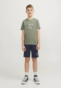 Zaļš īsroku T-krekls ar stilizētu logotipu, kopā ar tumši ziliem šortiem un melnām sporta kurpēm. Zeķes ir baltas ar logotipa druku aprocē.