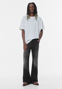 T-shirt oversize grigio chiaro con scollatura rotonda, abbinato a jeans svasati scuri. Il modello indossa scarpe slip-on marroni e gioielli in metallo.