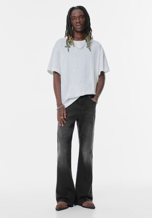 Lichtgrijze oversized t-shirt met een ronde hals, gecombineerd met donkere, uitlopende jeans. Het model draagt een paar bruine instap schoenen en metalen sieraden.