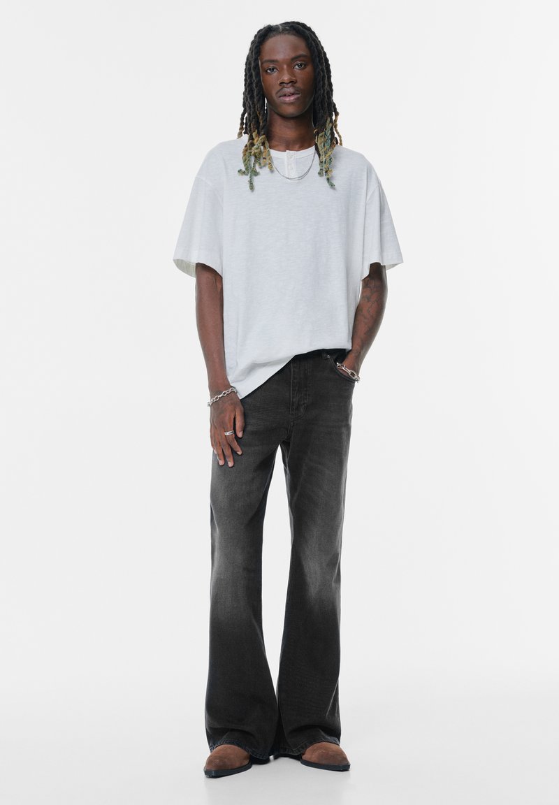 T-shirt oversize grigio chiaro con scollatura rotonda, abbinato a jeans svasati scuri. Il modello indossa scarpe slip-on marroni e gioielli in metallo.