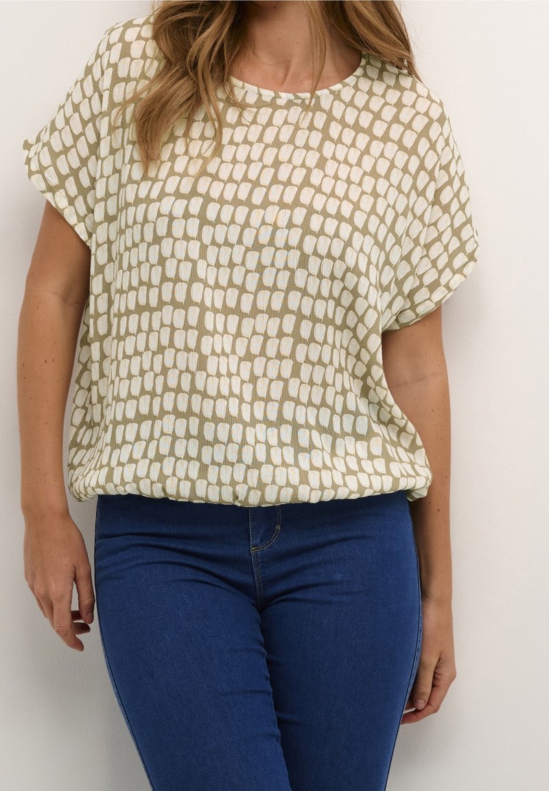 Femme portant un chemisier à manches courtes beige et blanc à motifs, rentré dans un jean bleu foncé ajusté, sur un fond uni.