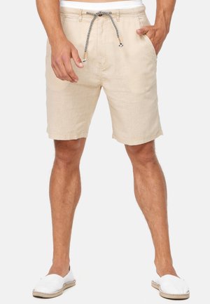 Beige Leinen-Shorts mit einem Tunnelzugbund, die eine dezente Textur und eine entspannte Passform aufweisen. Kombiniert mit weißen Slip-On-Schuhen.