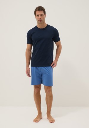 Man staat op blote voeten, draagt een marineblauw t-shirt en lichtblauwe korte broek tegen een effen achtergrond.