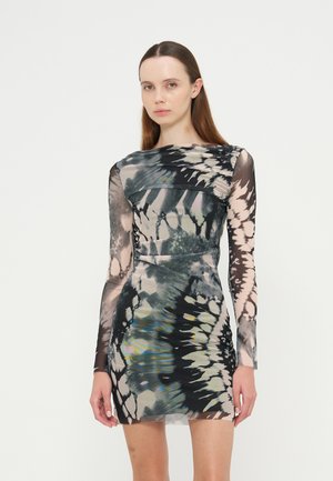 Jeune femme portant une mini-robe ajustée à manches longues avec un motif abstrait tie-dye noir, gris et beige, debout devant un fond uni.