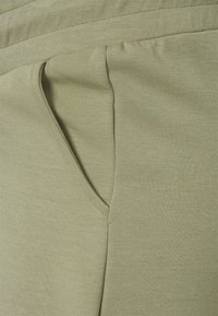 Tecido verde claro com uma textura suave e macia, apresentando um bolso lateral. O material parece elástico e confortável para uso casual.