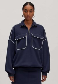 Marineblauwe pullover met een ritssluiting bij de kraag, twee grote voorzakken, witte piping accenten en losse mouwen. Gemaakt van zachte stof.