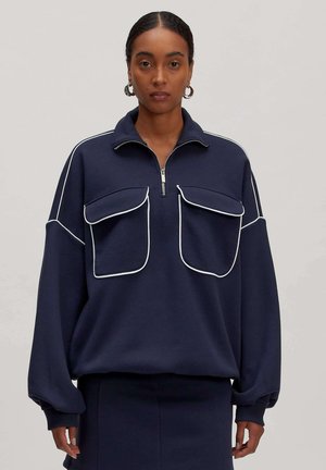 Marineblauer Pullover mit Reißverschlusskragen, zwei großen Fronttaschen, weißen Paspelierungen und locker sitzenden Ärmeln. Hergestellt aus einem weichen Stoff.