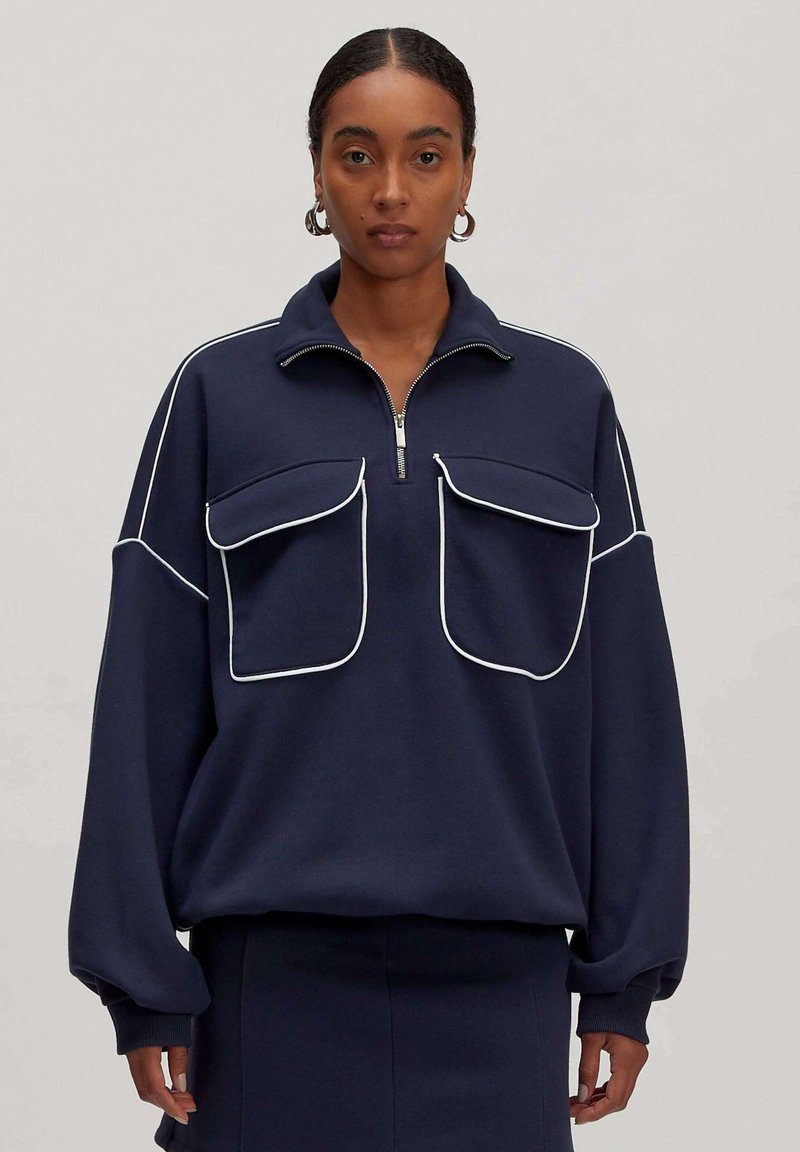 Marineblauwe pullover met een ritssluiting bij de kraag, twee grote voorzakken, witte piping accenten en losse mouwen. Gemaakt van zachte stof.