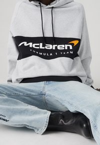 Szara bluza z kapturem z czarnym panelem, na którym znajduje się logo "McLaren Formula 1 Team" w białym i pomarańczowym kolorze. Uzupełniona jasnoniebieskimi dżinsami i czarnymi butami.