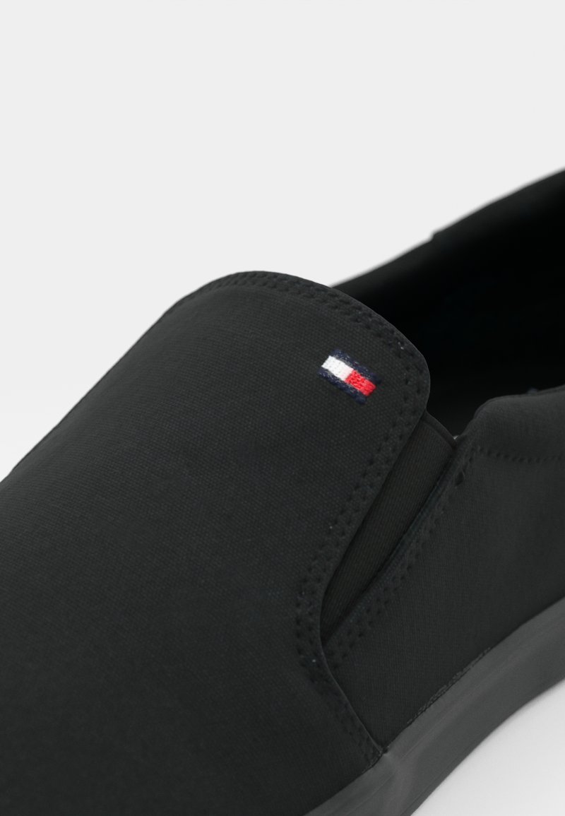 Chaussure noire à enfiler en toile, avec des panneaux latéraux élastiques et un petit emblème en rouge, blanc et bleu près de l'ouverture.